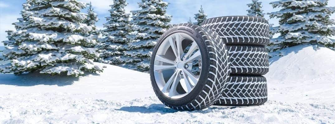 Winter tires stacked on snow-covered ground with snow-laden evergreen trees.