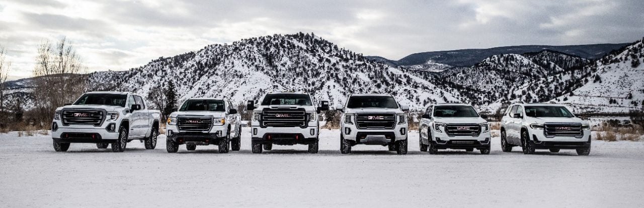 Six GMC trucks lined up on snowy terrain, with snow-covered mountains in the background.